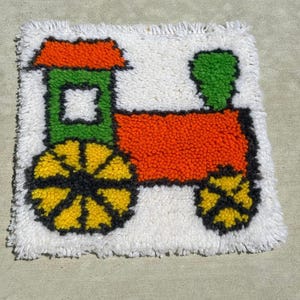 Puede incluir: Una colorida alfombra hecha a mano con un diseño de tren. El tren tiene un cuerpo naranja, cabina verde y ruedas amarillas con detalles negros. El fondo es blanco y la alfombra tiene un aspecto texturizado y afelpado.