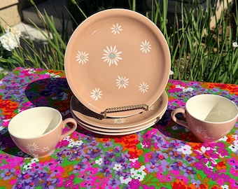 May include: A set of dusty pink ceramic dinnerware featuring a white daisy pattern with light blue centers. The set includes a stack of four round plates and two matching tea cups with handles, all displaying a consistent floral motif throughout.