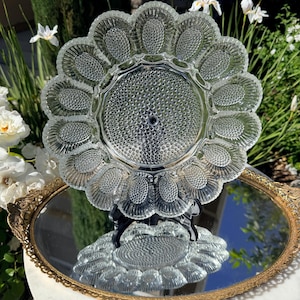 May include: A clear glass oyster serving platter with a scalloped edge and individual compartments. The platter is displayed on a mirrored tray with a gold-colored ornate frame, reflecting the surrounding greenery and flowers.