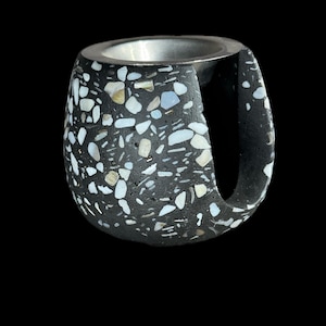 May include: A rounded, black vessel featuring a speckled terrazzo pattern with white and light-colored fragments embedded throughout. The object has a smooth, metallic silver-toned interior rim and a large, vertical cutout on the right side.