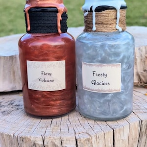 May include: Two glass jars with labels and decorative lids. The jar on the left is filled with a red liquid and has a black lid with a copper top. The label reads "Fiery Volcano". The jar on the right is filled with a blue liquid and has a brown lid with a white top. The label reads "Frosty Glaciers".