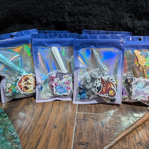 May include: Four clear plastic bags with iridescent holographic lining, each containing a variety of dice, miniature figurines, and stickers. The bags are labeled "Roll for" and feature a variety of fantasy-themed designs.