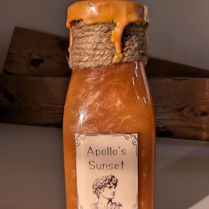 May include: A glass bottle with a gold glitter finish and a label that reads "Apollo's Sunset". The bottle is topped with a brown rope and a layer of orange wax.