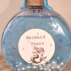 May include: A round glass bottle filled with blue liquid and iridescent glitter. The bottle has a cork stopper sealed with blue wax. A white label with the words "Mermaid Tears" and a mermaid illustration is affixed to the bottle. The bottle is resting on a bed of sand.