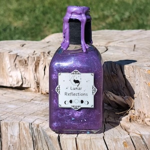 May include: A small glass bottle containing a purple glitter liquid. The bottle has a black wax seal on the top and a label that reads "Lunar Reflections" with a crescent moon and a cat design.