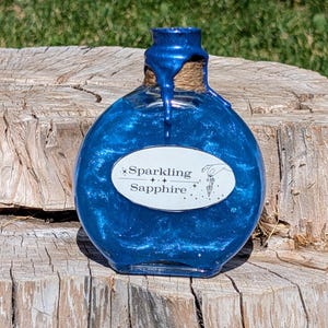 May include: A small, round glass bottle containing a blue, glittery liquid. The bottle has a cork stopper and a white label that reads "Sparkling Sapphire".