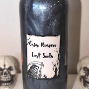 May include: A dark, shimmering bottle with a black cap and a label that reads "Grim Reapers Lost Souls." The label features a Grim Reaper illustration. The bottle is surrounded by two skull decorations, suggesting a Halloween or gothic theme.