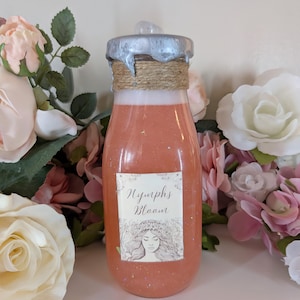 May include: A clear glass bottle with a silver lid and a brown twine accent. The bottle is filled with a pink liquid with glitter. The label on the bottle reads "Nymphs Bloom" and features a drawing of a woman with flowers in her hair.