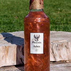 May include: A glass bottle with a copper-colored liquid inside. The bottle is sealed with a brown wax seal and has a label that reads "Phoenix Ashes".