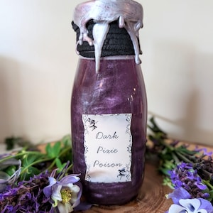 May include: A purple glass bottle with a silver-colored, dripped wax seal. The label reads "Dark Pixie Poison" with fairy illustrations. The bottle is surrounded by purple and white flowers and greenery.