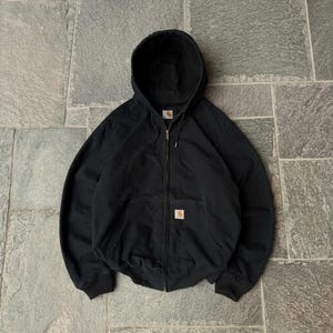 Pode incluir: Uma camisola preta com capuz e fecho de correr integral, capuz com cordão e dois bolsos frontais. Uma pequena etiqueta retangular da marca Carhartt está cosida na zona do bolso frontal esquerdo da peça de vestuário.