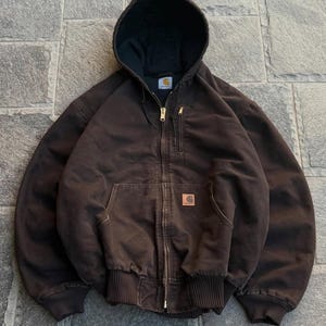Pode incluir: Um casaco com capuz e fecho de correr em castanho escuro, com forro interior preto, um bolso vertical com fecho no peito e um bolso frontal tipo canguru. Um emblema quadrado da marca Carhartt está cosido no bolso frontal do casaco.
