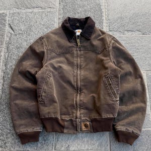 May include: A brown Carhartt jacket featuring a dark brown corduroy collar, a full-length front zip, two slanted side pockets, and a ribbed knit hem and cuffs. A small rectangular Carhartt brand patch is stitched onto the lower front left.