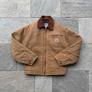 Könnte beinhalten: Eine beigefarbene Canvas-Jacke mit braunem Cordkragen, durchgehendem Frontreißverschluss und einer Brusttasche mit Reißverschluss. Ein quadratischer Carhartt-Markenaufnäher ist auf der linken Brustseite aufgenäht und die Ärmel haben geknöpfte Manschetten.