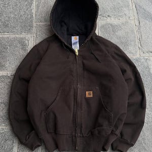 May include: A dark brown zip-up hooded jacket featuring a black interior lining, a brass-coloured metal zip, and a square Carhartt brand patch on the front kangaroo pocket. The garment includes ribbed cuffs and a ribbed hem for a secure fit.