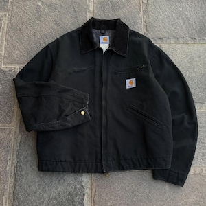Puede incluir: Una chaqueta negra Carhartt con cuello de pana oscuro, cremallera frontal completa y mangas largas con puños ajustables de botón. La prenda incluye un bolsillo en el pecho con cremallera y un parche cuadrado con el logotipo de Carhartt.