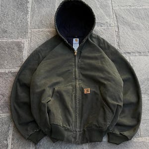 Può includere: Una giacca con cappuccio e zip color verde oliva scuro, dotata di chiusura frontale con cerniera, cappuccio con coulisse e fodera interna scura, polsini e orlo a coste, e un'etichetta quadrata del marchio Carhartt sulla tasca a marsupio.