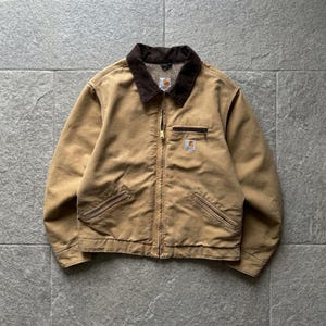 Op de afbeelding: Een beige canvas jas met een donkerbruine corduroy kraag, een volledige ritssluiting aan de voorzijde en twee schuine zakken onderaan. Aan de linkerzijde bevindt zich een kleine borstzak met ritssluiting en een rechthoekig Carhartt-label.