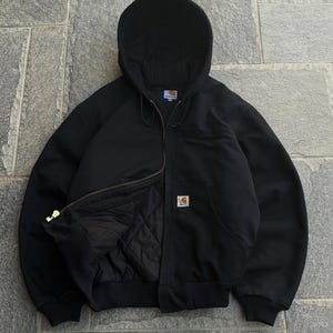 Op de afbeelding: Een zwarte jas met capuchon, een ritssluiting over de hele lengte, geribbelde boorden en zoom, en een gewatteerde voering. De jas is voorzien van een Carhartt-merklabel op de voorzak en een klein merklabel bij de binnenhals.
