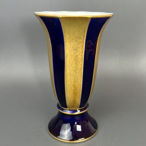 Op de afbeelding: Een decoratieve vaas met een uitlopende bovenkant en een voetstuk. De vaas heeft verticale gouden en marineblauwe strepen, met gouden rand langs de boven- en onderkant. De binnenkant is wit.