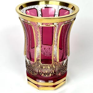 Puede incluir: Un jarrón de cristal decorativo con cuerpo rojo rubí y detalles dorados. El jarrón presenta un diseño geométrico con ribete dorado alrededor del borde y la base. Intrincados patrones dorados adornan la parte inferior del jarrón.