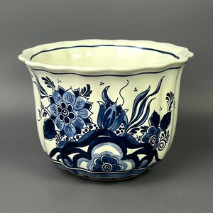 Może przedstawiać: Ceramiczna doniczka z falistym brzegiem, posiadająca kremową podstawę ozdobioną misternymi ciemnoniebieskimi wzorami kwiatowymi i stylizowanymi motywami botanicznymi. Naczynie ma zaokrąglony, żłobkowany korpus z gładkim, błyszczącym wykończeniem.