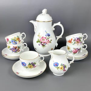 May include: A white porcelain tea set with floral patterns. The set includes a teapot, creamer, cups, and saucers. The floral design features pink, blue, yellow, and purple flowers with gold trim. The set is arranged on a grey surface.