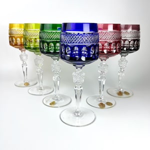 Puede incluir: Seis copas de vino de cristal de colores. Cada copa presenta un cuenco de diferente color, incluyendo amarillo, verde, azul y rojo. Los tallos son de cristal transparente con una base decorativa. Las copas están dispuestas en fila sobre una superficie blanca.