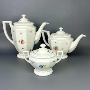 Può includere: Un servizio da tè in porcellana bianca con decorazioni floreali. Il set comprende una teiera, una teiera più piccola e una zuccheriera. Ogni pezzo ha un bordo dorato ed è decorato con fiori colorati. Il set è presentato su sfondo grigio.