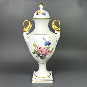 Pode incluir: Uma urna de porcelana branca com tampa, apresentando buquês de flores pintados à mão em rosa, azul e amarelo. O vaso inclui duas alças douradas em forma de cisne e detalhes em ouro na base, na borda e no topo da tampa sobre um pedestal.