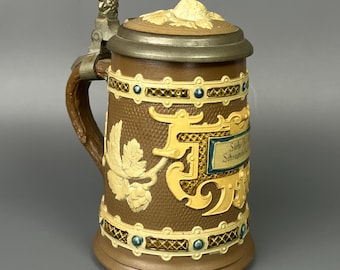 May include: A brown ceramic beer stein with a decorative design. The stein features a hinged lid, a handle, and ornate relief carvings. The design includes a banner with text, leaf motifs, and blue and yellow accents. The metalwork is silver-toned.