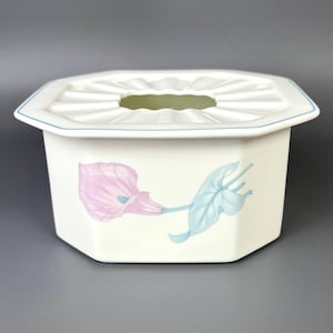 May include: An octagonal white ceramic tissue box with a floral design. It features pink calla lilies and blue-green leaves. The lid has a central opening and a light blue trim.