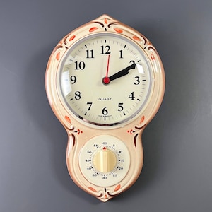 Puede incluir: Reloj de pared de cuarzo color crema con temporizador integrado. La esfera del reloj tiene números y manecillas negras, con un segundero rojo. El temporizador tiene un dial dorado y una escala de 0 a 60 minutos. El reloj está enmarcado con un borde decorativo color melocotón.