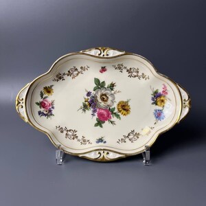 Op de afbeelding: Een wit ovaal serveerblad met een gouden rand en een bloemenmotief. Het blad heeft een centraal boeket van roze, witte en gele bloemen met groene bladeren. De rand is versierd met een herhalend patroon van gouden bloemen en bladeren.