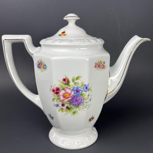 Puede incluir: Tetera de porcelana blanca con un diseño floral. La tetera tiene un borde dorado alrededor del pico y el asa. Las flores son rosas, azules, amarillas y moradas.