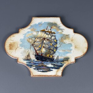 May include: A decorative ceramic tile with a quatrefoil shape featuring a painted scene of a large sailing ship on a turbulent blue ocean under a cloudy sky. The surface has a visible crackled finish with cream-colored edges and brown trim.