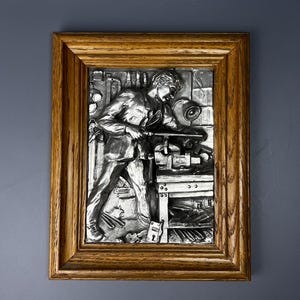 Può includere: Una scultura da parete incorniciata in argento che raffigura una persona che lavora su un oggetto di metallo in un laboratorio. La scultura è dettagliata con strumenti e altri oggetti del laboratorio.