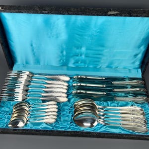 Könnte beinhalten: Ein komplettes Besteckset, einschließlich Gabeln, Messern und Löffeln, ordentlich in einem schwarzen Koffer mit türkisfarbenem Satinfutter angeordnet. Das Besteck hat ein dekoratives Muster auf den Griffen. Der Koffer ist geöffnet und zeigt das komplette Set.