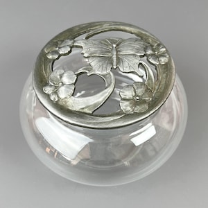 Könnte beinhalten: Ein klares Glasgefäß mit einem dekorativen Metalldeckel. Der Deckel zeigt ein Schmetterlings- und Blumendesign in Silber. Das Gefäß hat eine runde Form und steht auf einem neutralen Hintergrund. Das Gefäß wird wahrscheinlich zur Aufbewahrung oder Präsentation verwendet.