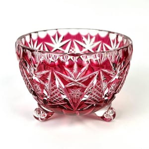 Peut inclure: Un bol en cristal taillé rouge rubis avec un motif d'éclat d'étoile. Le bol a une forme arrondie et repose sur trois petits pieds arrondis. Le design en cristal taillé crée un jeu d'ombre et de lumière, améliorant son attrait décoratif.