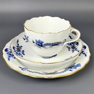 Könnte beinhalten: Ein weißes Porzellan-Teeservice, bestehend aus einer Tasse, einer Untertasse und einem Teller. Das Set hat ein blaues Drachen- und Blumenmuster mit goldenem Rand. Die Tasse hat einen gewellten Rand.