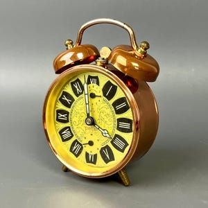 Könnte beinhalten: Ein Vintage-Wecker in Kupferoptik mit gelbem Zifferblatt und schwarzen römischen Ziffern. Die Uhr hat ein dekoratives Zifferblattdesign und einen goldfarbenen Griff und Glocken. Auf dem Zifferblatt der Uhr steht "peter".