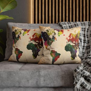 May include: Two square throw pillows with a world map design. The map is rendered in a watercolor style with vibrant colors, including red, green, yellow, and blue. The pillows are on a gray sofa.