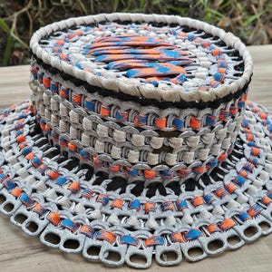 May include: A handmade hat constructed from recycled aluminum can tabs. The hat features a wide brim and a cylindrical crown, with a color scheme of gray, black, white, orange, and blue. The hat is a unique, eco-friendly accessory.