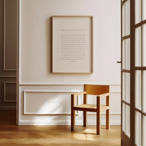May include: A framed print with a beige background and black text. The text reads "Philippians 2:1-2" and a passage from the Bible. The print is hanging on a white wall with a wooden chair in front of it.