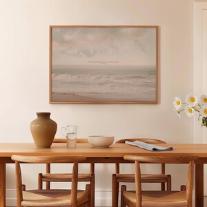 May include: A framed print of a seascape with a quote in the sky, hanging above a wooden dining table. The table is set with a vase, pitcher, bowl, and chairs. The print features a muted color palette of beige, gray, and blue.