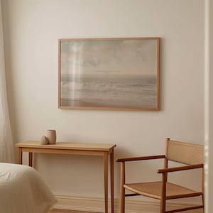 May include: A framed print of a seascape hangs on a cream-colored wall. The print features a muted color palette of beige, gray, and white, with text at the top. A wooden console table and chair are below the print, with a vase and decorative stone on the table.