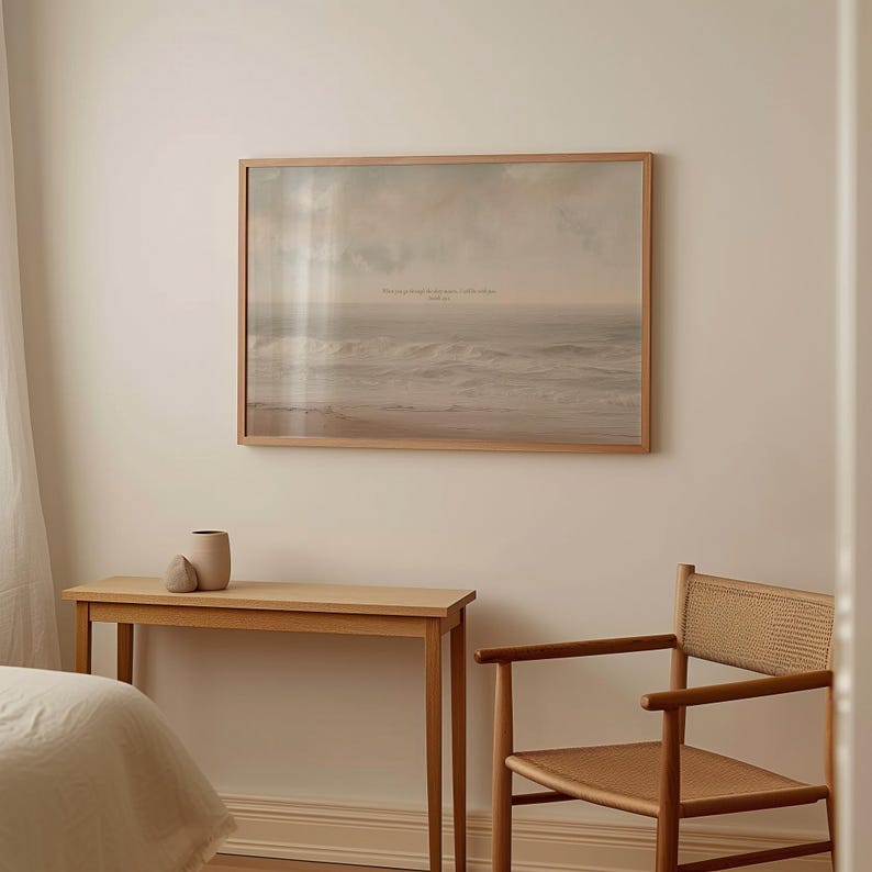 May include: A framed print of a seascape hangs on a cream-colored wall. The print features a muted color palette of beige, gray, and white, with text at the top. A wooden console table and chair are below the print, with a vase and decorative stone on the table.
