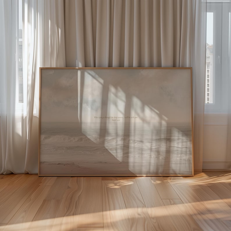 May include: A framed print featuring a serene ocean scene in muted tones. The artwork displays a seascape with gentle waves and a cloudy sky. Text at the bottom reads "When you go through the deep waters, I will be with you." The frame is a light wood color.
