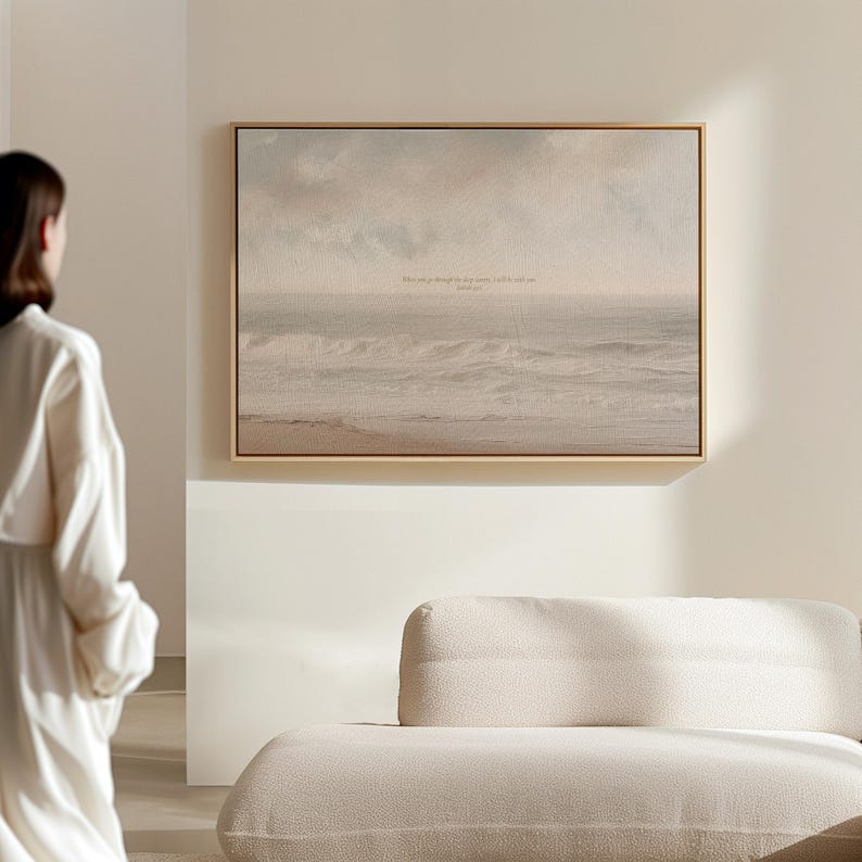 May include: A framed canvas print depicting a seascape with a muted color palette of beige, gray, and white. The artwork features a calm ocean under a cloudy sky, with a subtle text inscription. The frame is a light wood color.